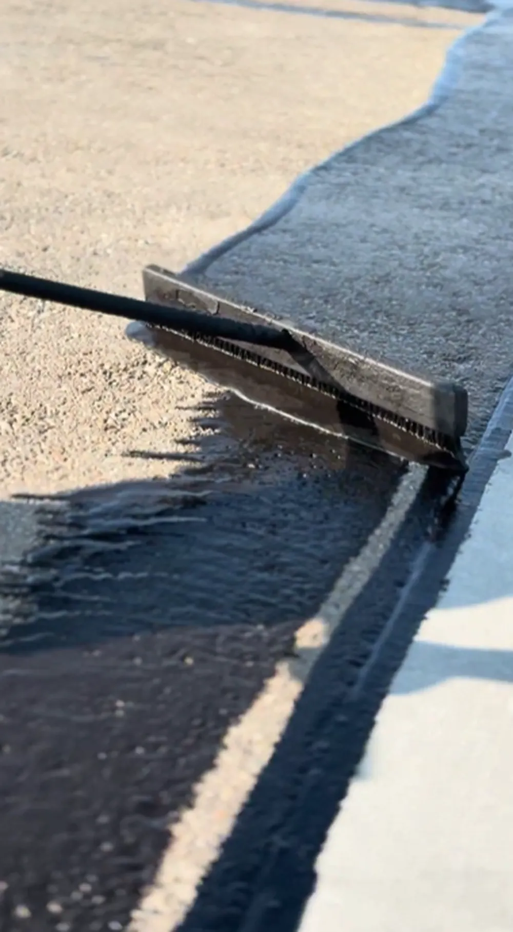 Macro close-up of a sealcoating squeegee brush spreading fresh black asphalt sealer along a concrete curb edge, showing the wet sealer texture and clean edge line on a parking lot in Nampa, Idaho