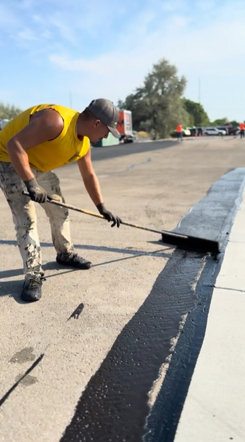 Superior Striping worker in yellow sleeveless shirt applying black asphalt sealcoating with a squeegee along a concrete curb edge on a commercial parking lot in Nampa, Idaho