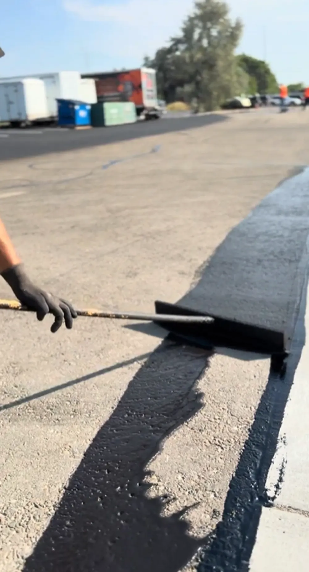 Worker wearing black gloves hand-applying asphalt sealcoating with a squeegee along a curb edge in a commercial parking lot in Nampa, Idaho