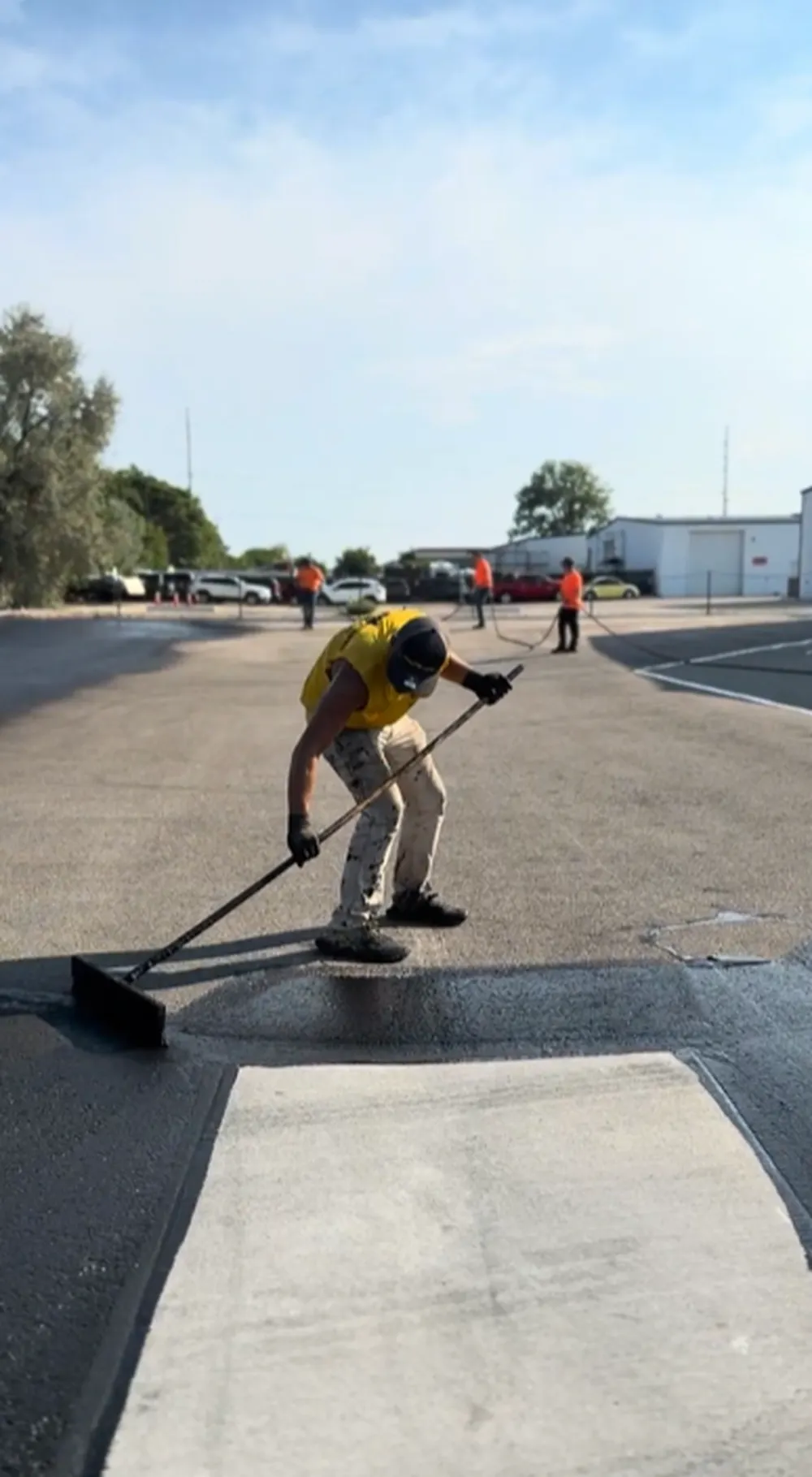 Superior Striping technician in yellow shirt pushing a squeegee to spread fresh asphalt sealcoating on a large commercial parking lot in Nampa, Idaho, with additional crew members and sealcoating equipment visible in the background