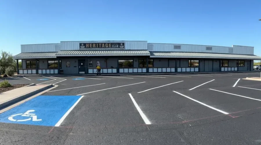 Freshly completed parking lot sealcoating and striping at Heritage Social Club, featuring bright white parking stall lines, a blue ADA handicap accessible space with wheelchair symbol, and a Superior Striping crew member visible near the building entrance under a clear blue sky.