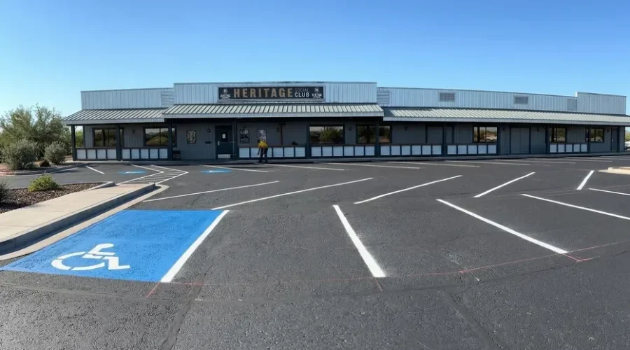 Freshly completed parking lot sealcoating and striping at Heritage Social Club, featuring bright white parking stall lines, a blue ADA handicap accessible space with wheelchair symbol, and a Superior Striping crew member visible near the building entrance under a clear blue sky.