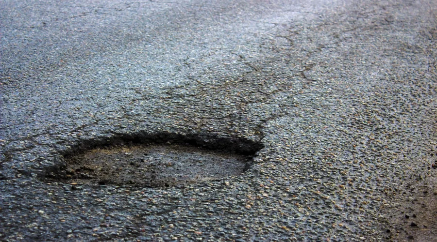 A close-up of a deep pothole on a worn asphalt driveway surface with visible cracks around it.