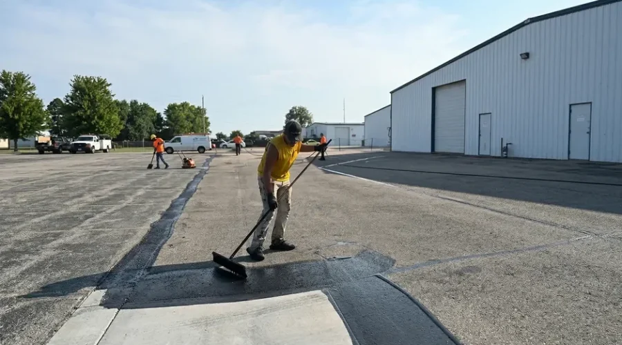 Wide-angle view of a full Superior Striping crew applying sealcoat across a large commercial asphalt parking lot in Nampa, Idaho, with one worker in a yellow shirt using a push broom in the foreground, additional crew members with equipment visible in the background, and a white industrial warehouse building on the right.