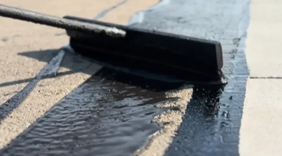 Extreme close-up ground-level shot of a squeegee spreading shiny wet asphalt sealcoating along a concrete curb edge on a commercial parking lot in Nampa, Idaho